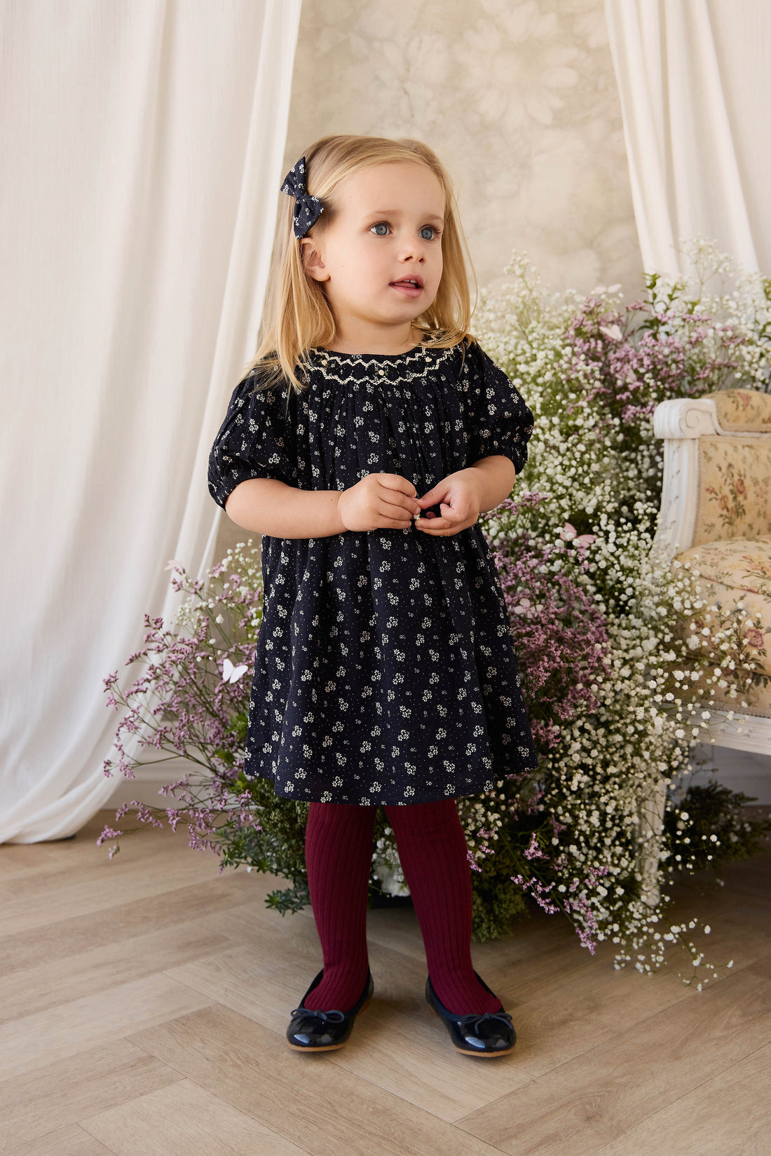 Young girl in a dark floral dress standing in front of white curtains and floral decorations.