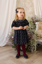 Young girl in a dark floral dress standing in front of white curtains and floral decorations.