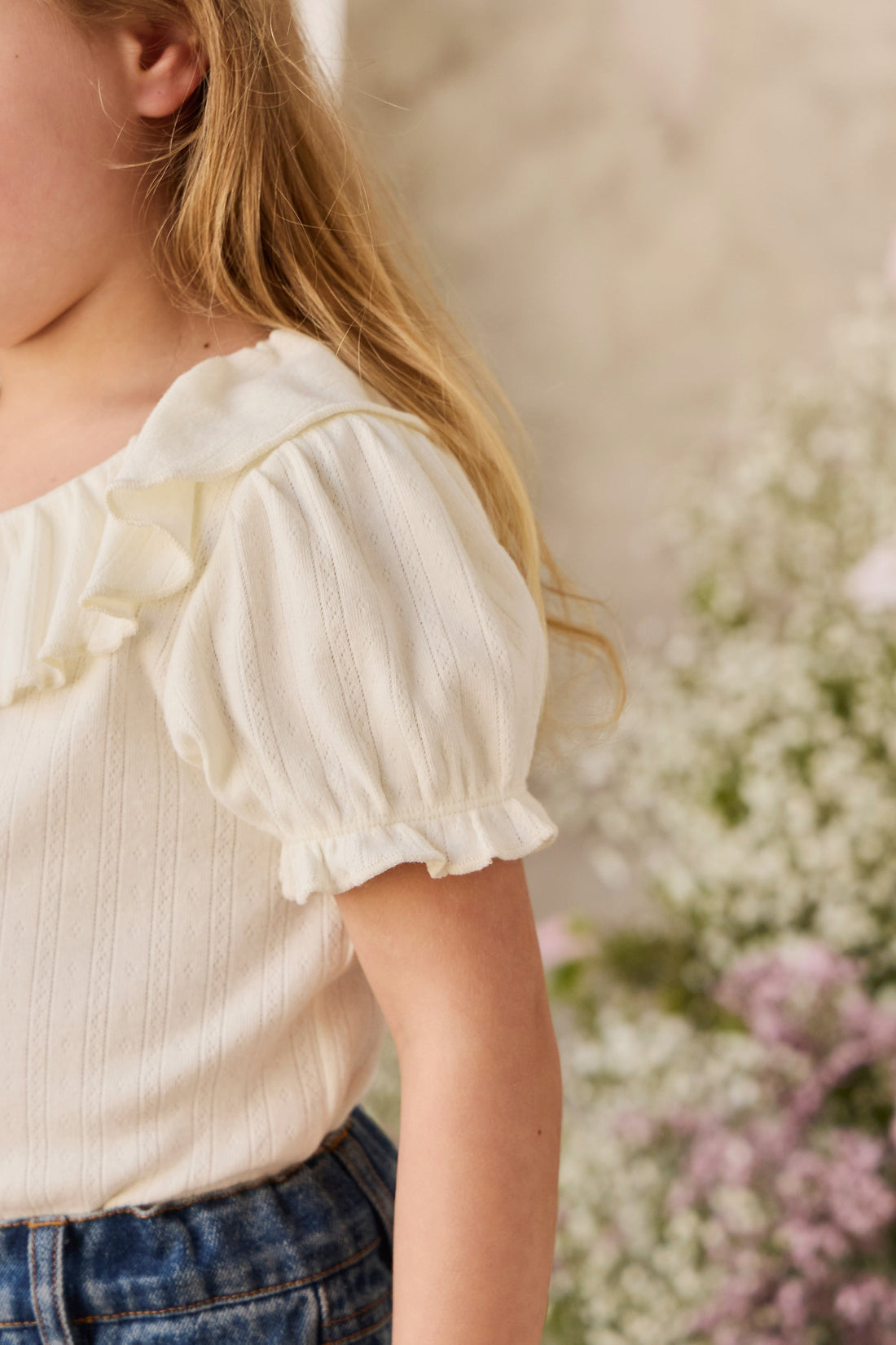 A close-up of a child wearing a cream-colored short-sleeve top with ruffled trim and puff sleeves, paired with blue jeans, set against a soft-focus background of white and pink flowers.