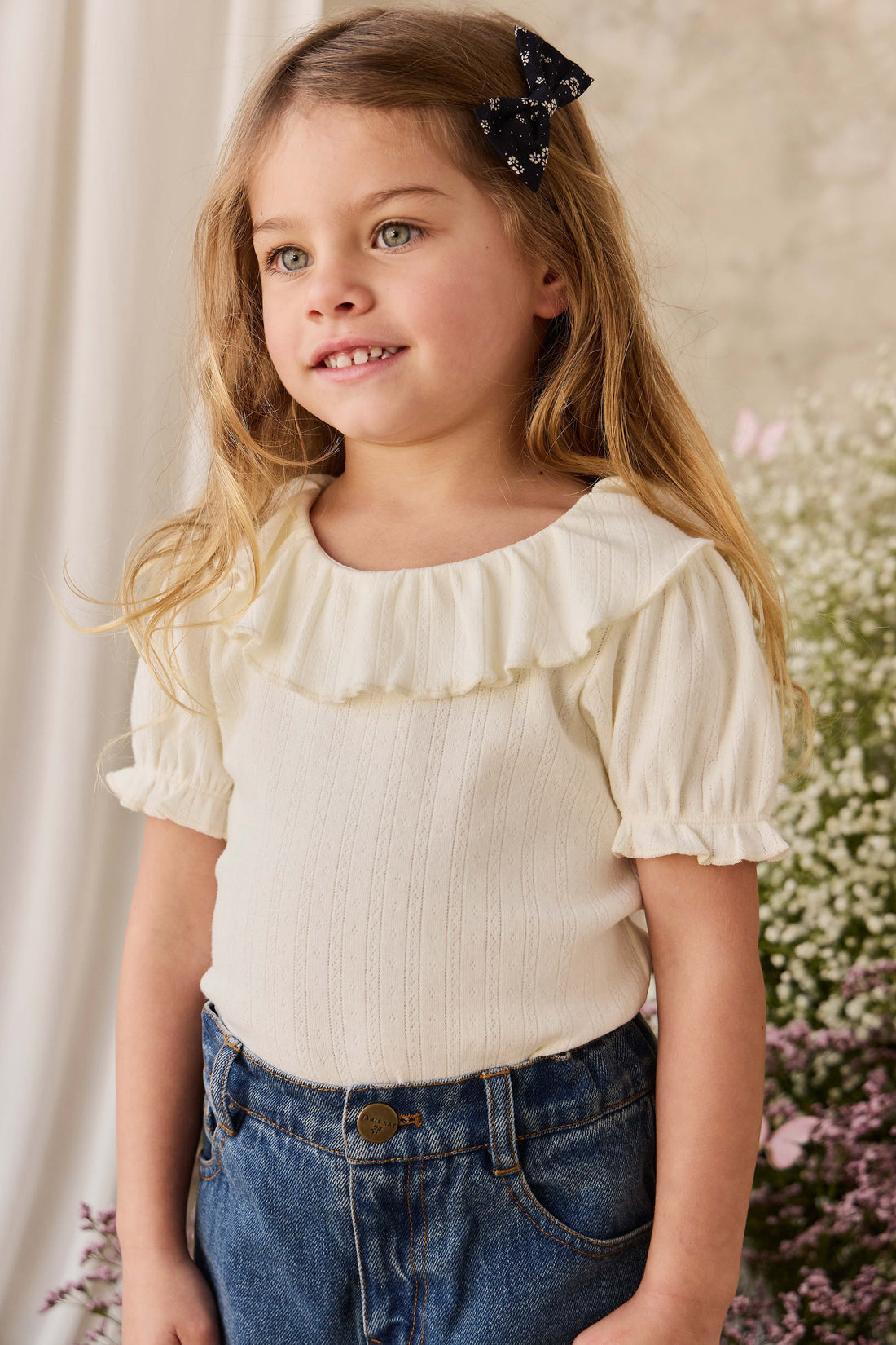 A young girl with blonde hair wearing a cream-colored short-sleeve top with a ruffled collar and blue jeans, standing in front of a soft beige background with flowers.