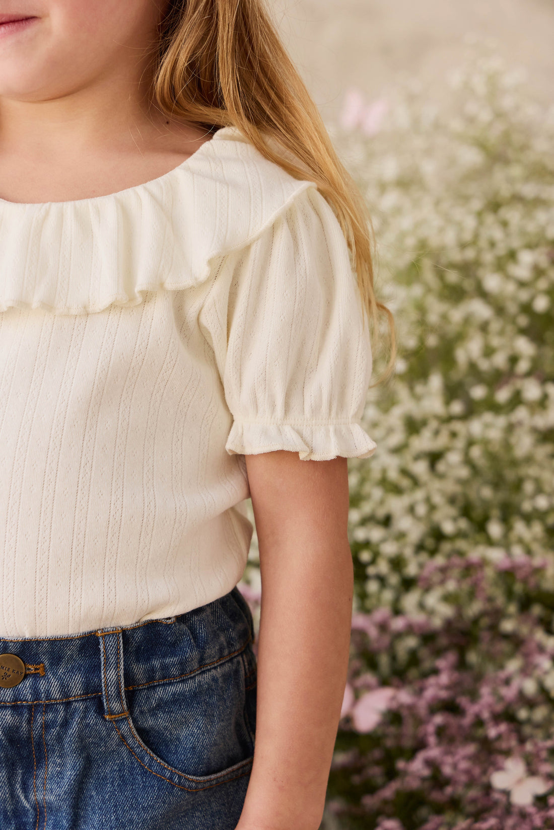 Child wearing a white blouse with ruffled sleeves and blue jeans standing in front of flowers.