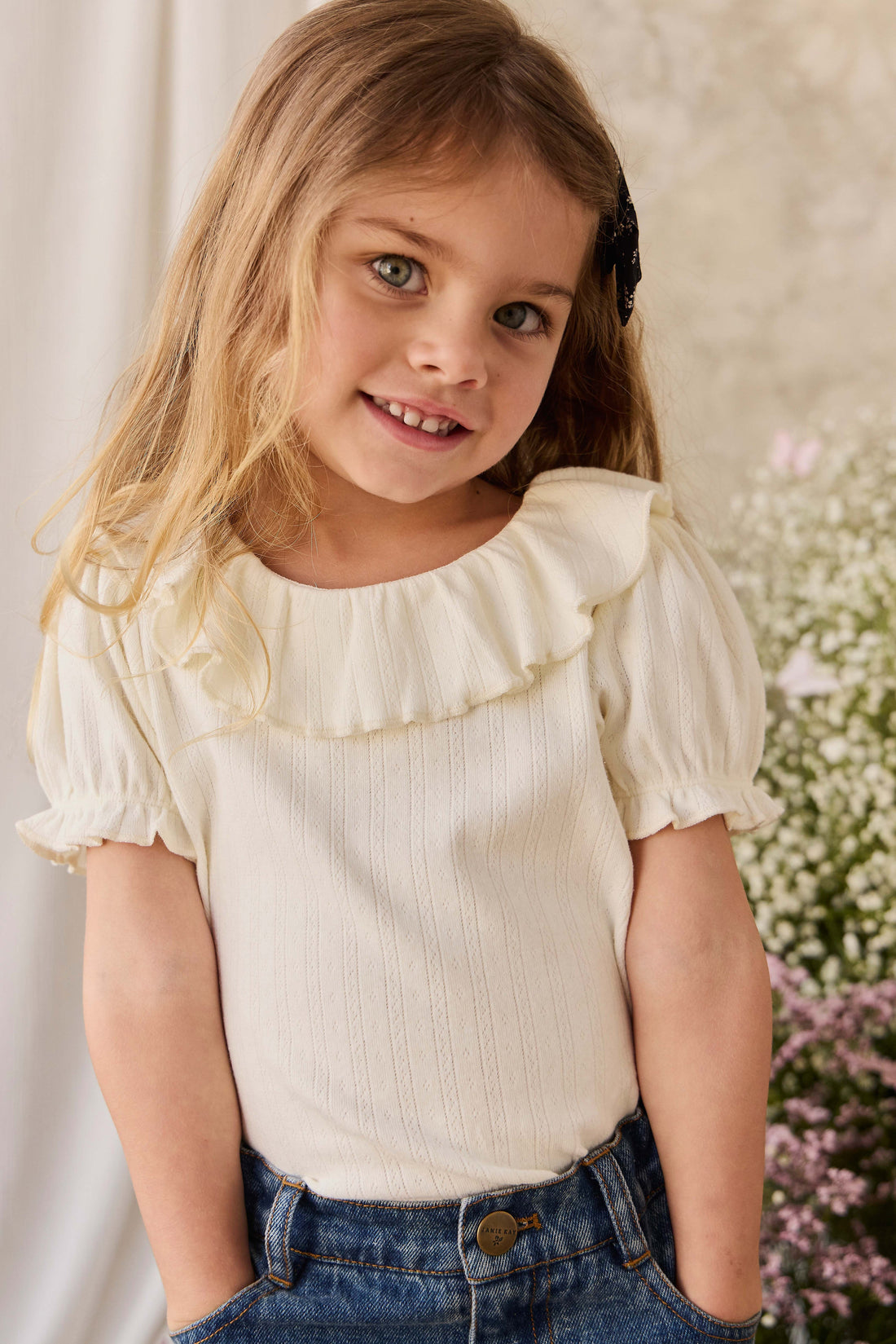Young girl wearing a white blouse with ruffled details and blue jeans, standing against a neutral background with flowers.