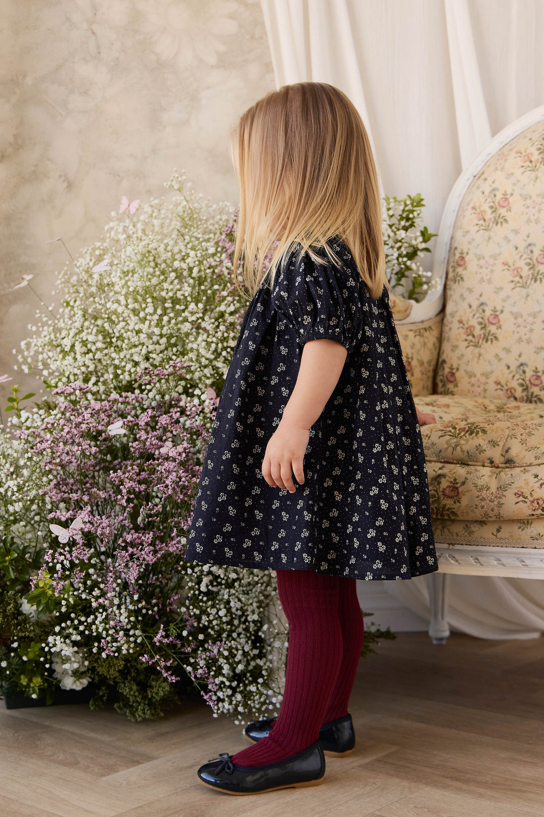 Young girl in a floral dress standing in a room with flowers and a sofa.