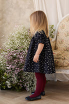 Young girl in a floral dress standing in a room with flowers and a sofa.