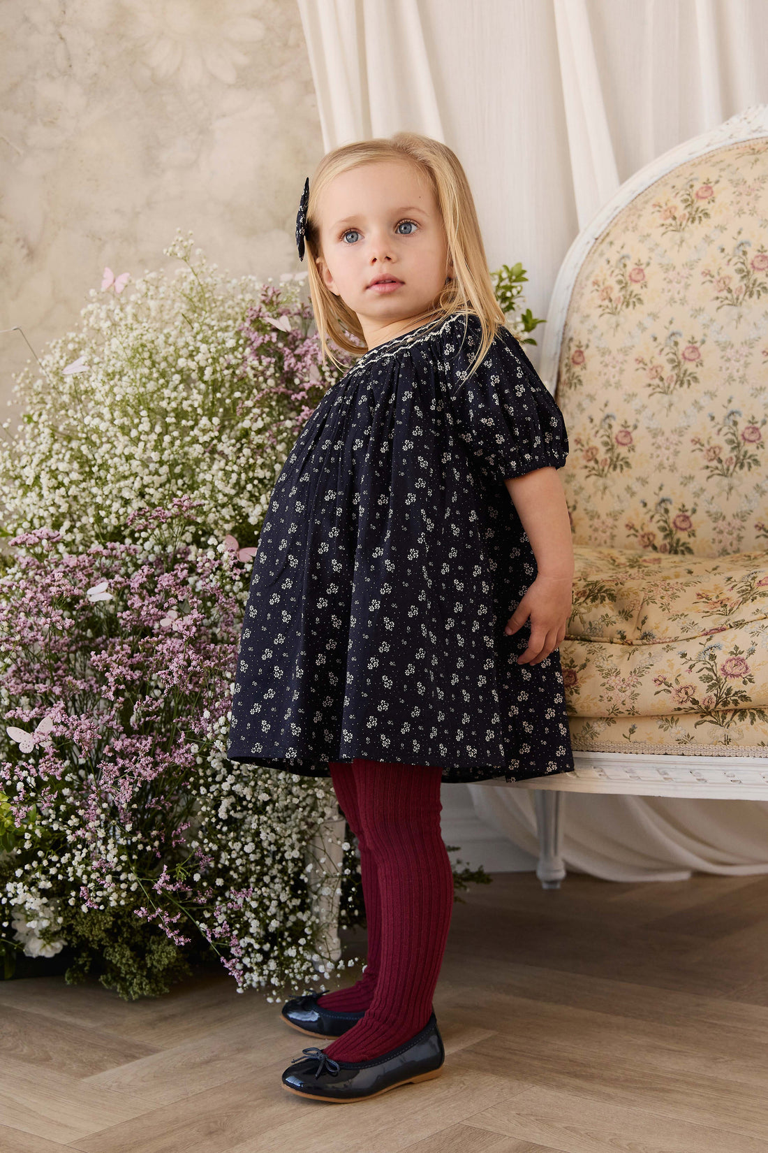 A young girl stands in a vintage-style room wearing a navy blue short-sleeve dress with a white floral pattern, burgundy ribbed tights, and dark navy shoes. She is positioned in front of a floral-patterned armchair and a large bouquet of white and pink flowers.