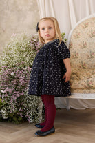 A young girl stands in a vintage-style room wearing a navy blue short-sleeve dress with a white floral pattern, burgundy ribbed tights, and dark navy shoes. She is positioned in front of a floral-patterned armchair and a large bouquet of white and pink flowers.