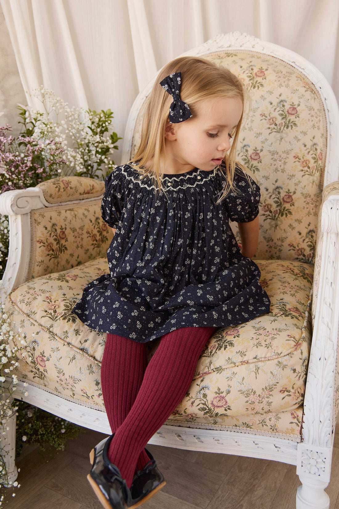 A young girl with blonde hair sits on a vintage floral armchair, wearing a navy blue floral dress, burgundy tights, and black shoes.