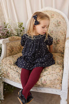 A young girl with blonde hair sits on a vintage floral armchair, wearing a navy blue floral dress, burgundy tights, and black shoes.