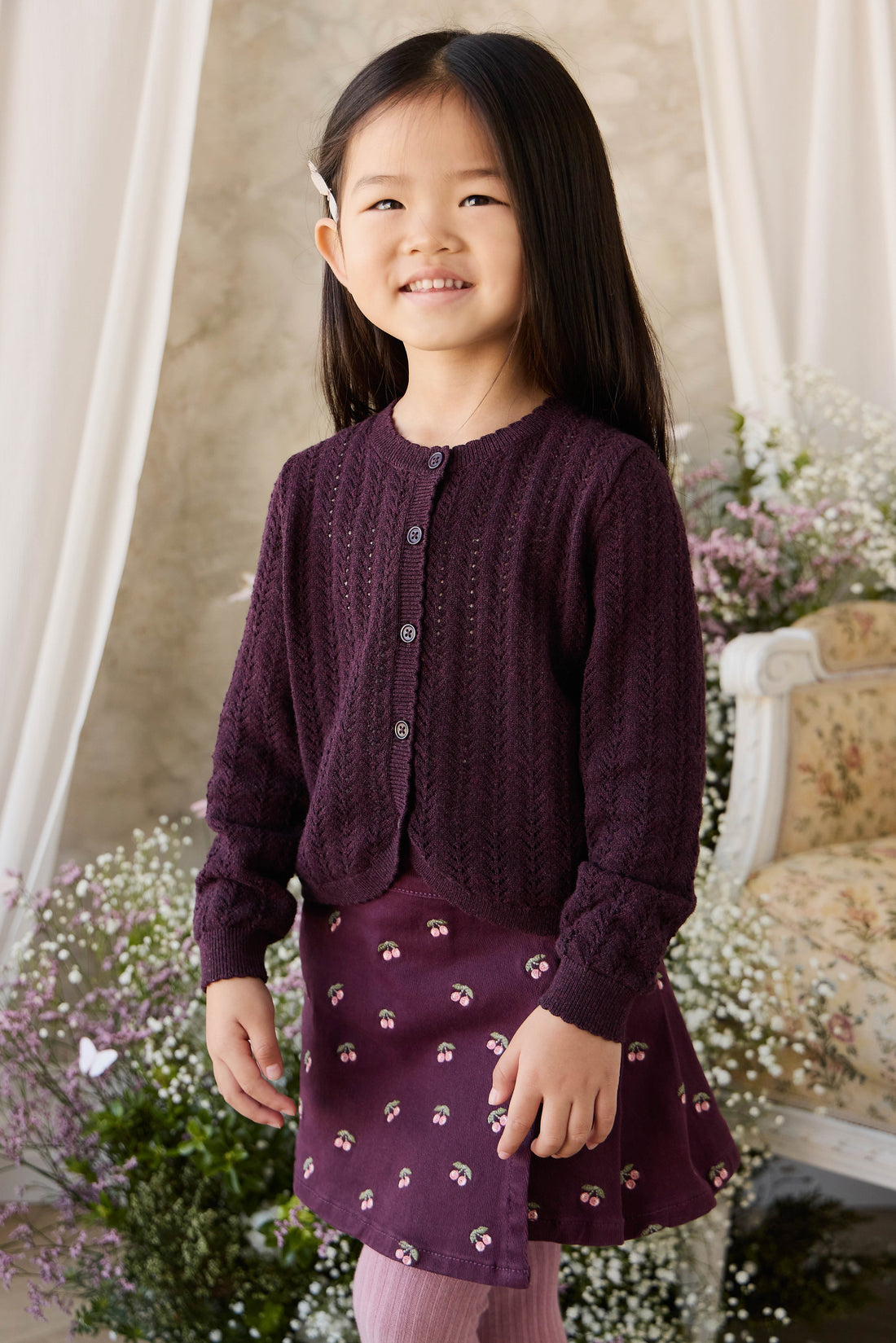 Young girl wearing a purple cardigan and skirt standing in a decorative room with flowers and a chair.