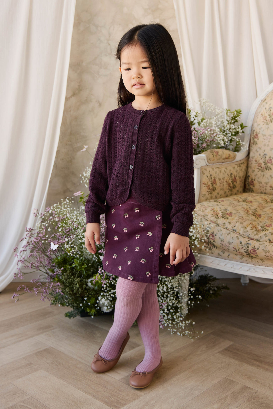 Young girl in a purple outfit standing in a decorated room with flowers and furniture.