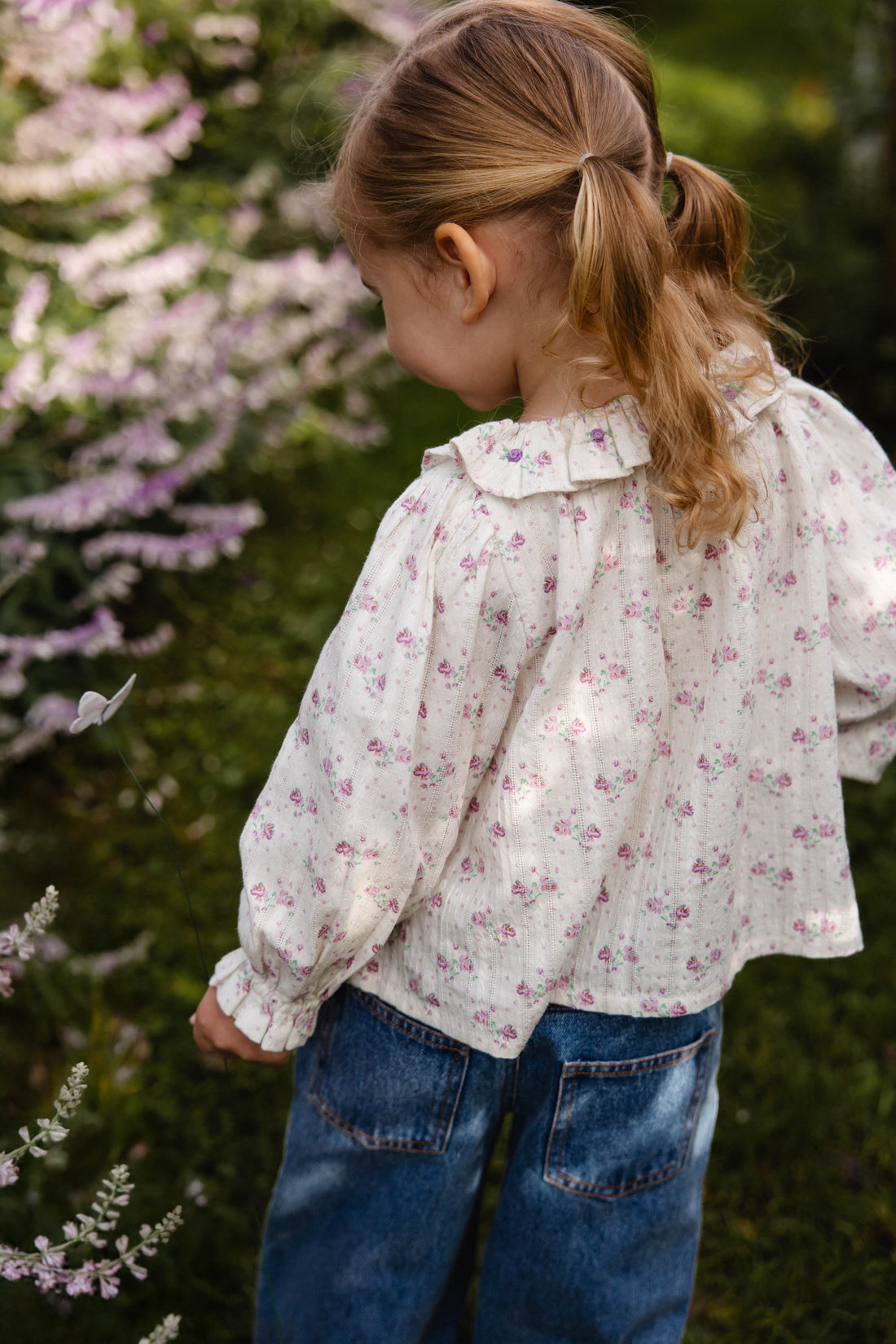 Child wearing a floral blouse and blue jeans standing in a garden with flowers.