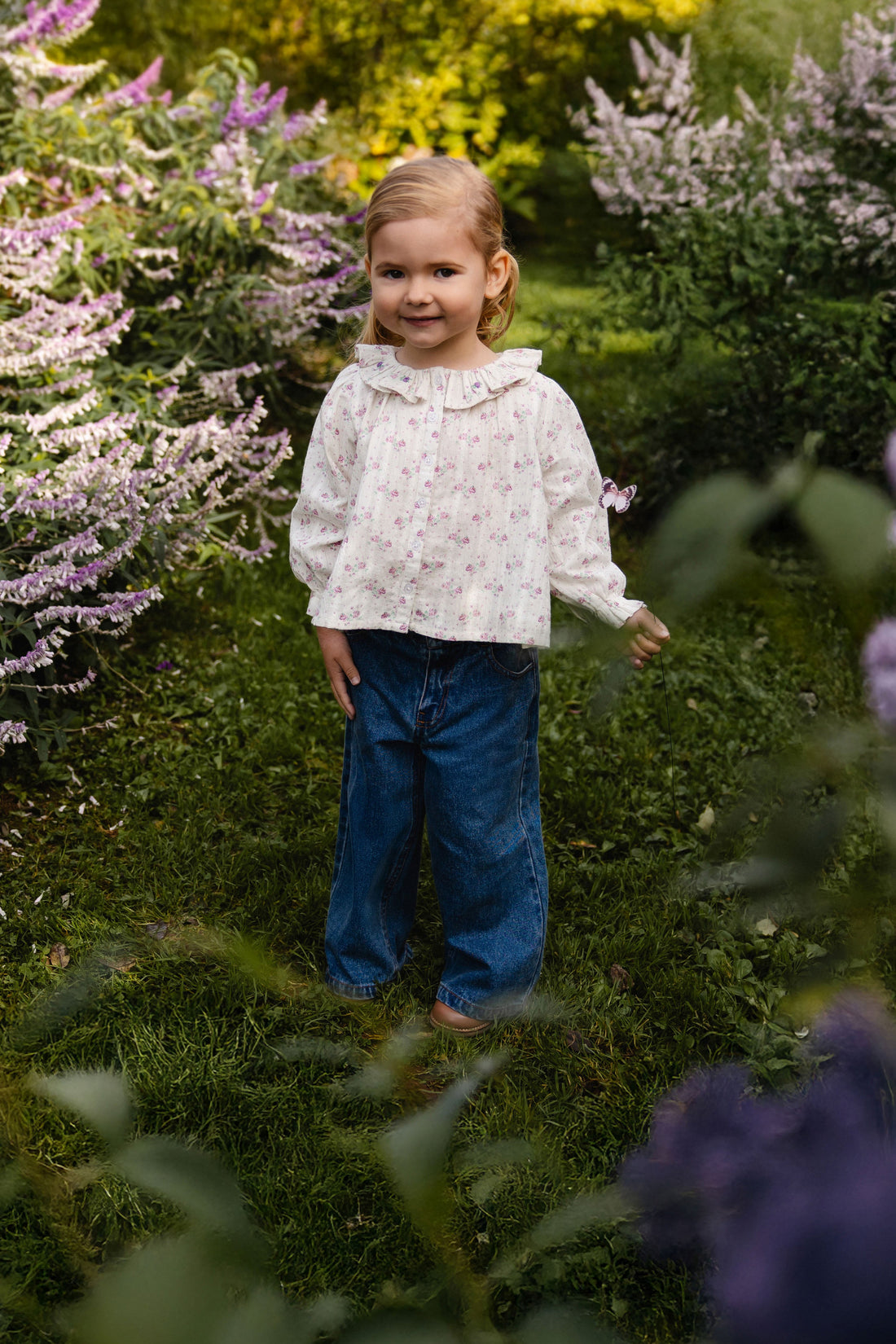 A young girl with blonde hair stands in a lush garden wearing a white long-sleeve blouse with a tiny floral print and blue jeans.