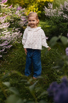 A young girl with blonde hair stands in a lush garden wearing a white long-sleeve blouse with a tiny floral print and blue jeans.