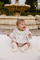 A baby girl sitting outdoors on a white blanket in front of a stone fountain, wearing a cream floral long-sleeve romper with a ruffled collar and matching cream knit booties.