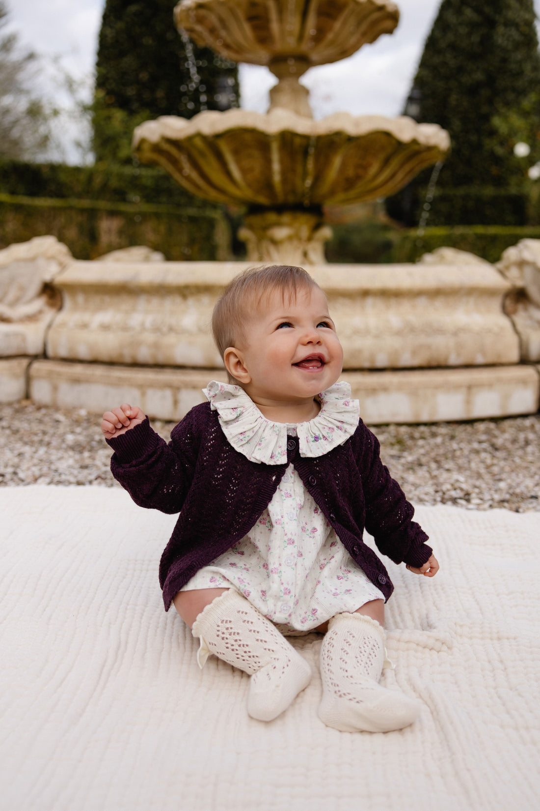 A baby girl sitting on a white textured blanket in front of a stone fountain, wearing a dark plum knit cardigan over a white floral dress with a ruffled collar and cream knit booties.