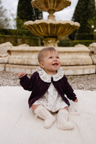 A baby girl sitting on a white textured blanket in front of a stone fountain, wearing a dark plum knit cardigan over a white floral dress with a ruffled collar and cream knit booties.