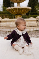 Baby in a purple cardigan and white dress sitting on a blanket with a fountain in the background
