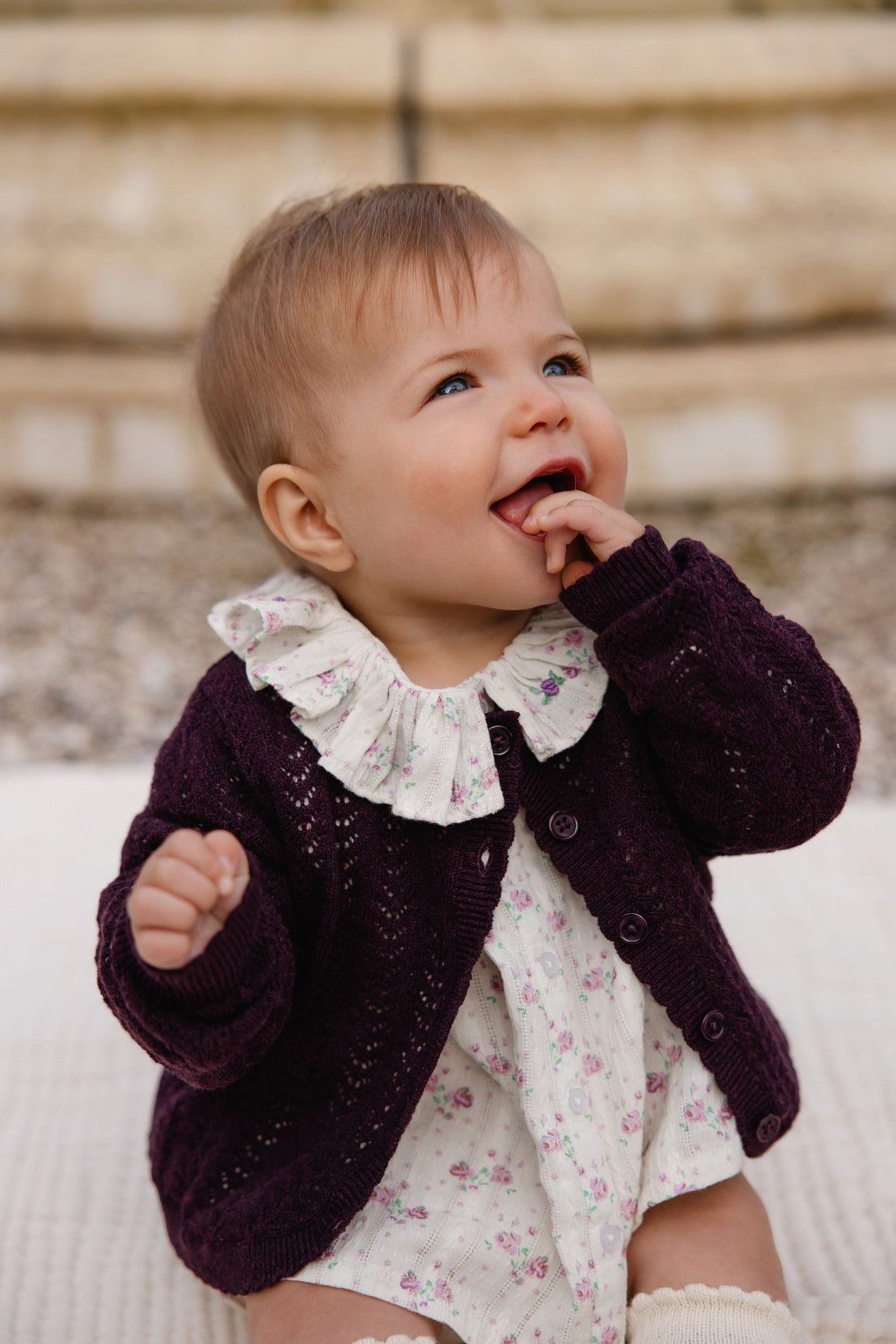 A baby girl wearing a dark plum purple knit cardigan over a white floral romper with a ruffled collar, sitting outdoors against a soft-focus stone wall background.