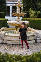 Child in a black dress standing in front of a stone fountain with greenery around.
