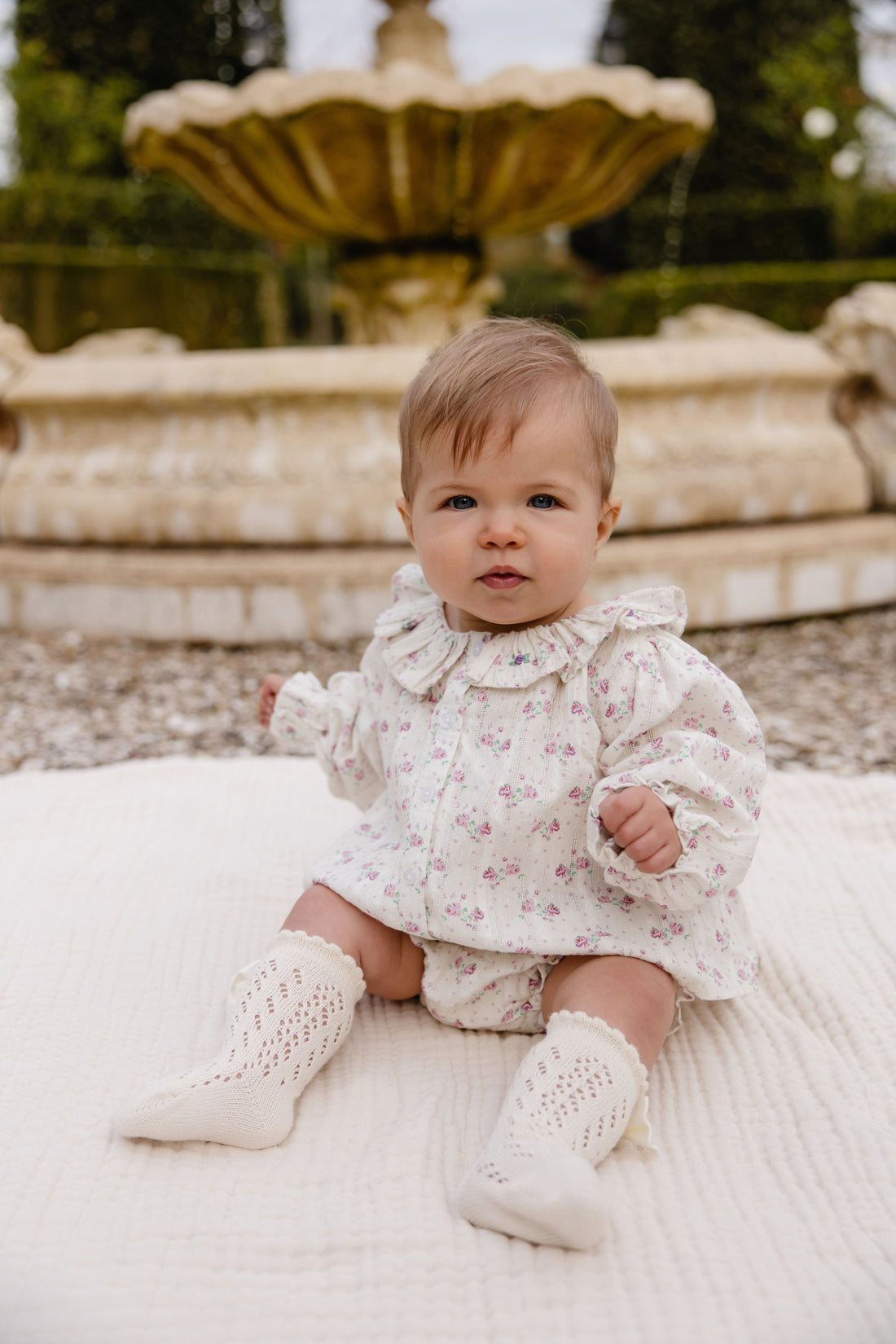A baby sitting outdoors in front of a stone fountain, wearing a cream floral romper with a ruffled collar and matching cream knit booties.