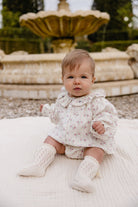A baby sitting outdoors in front of a stone fountain, wearing a cream floral romper with a ruffled collar and matching cream knit booties.