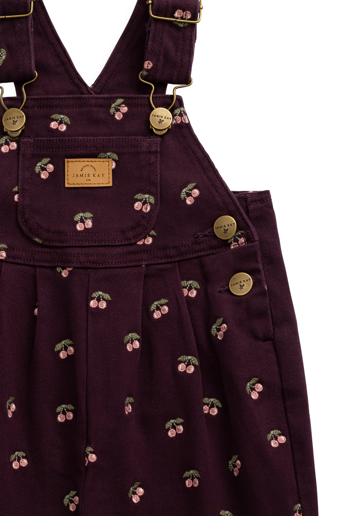 Close-up of a dark plum corduroy pinafore dress with a cherry print and gold-tone hardware, featuring a front patch pocket with a 'JAMIE KAY' label.