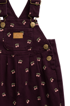Close-up of a dark plum corduroy pinafore dress with a cherry print and gold-tone hardware, featuring a front patch pocket with a 'JAMIE KAY' label.