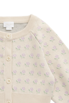 Close-up of a cream-colored knit cardigan sweater featuring a delicate all-over pattern of small purple flowers with green leaves, accented by a ribbed collar, cuffs, and hem, and a button closure.