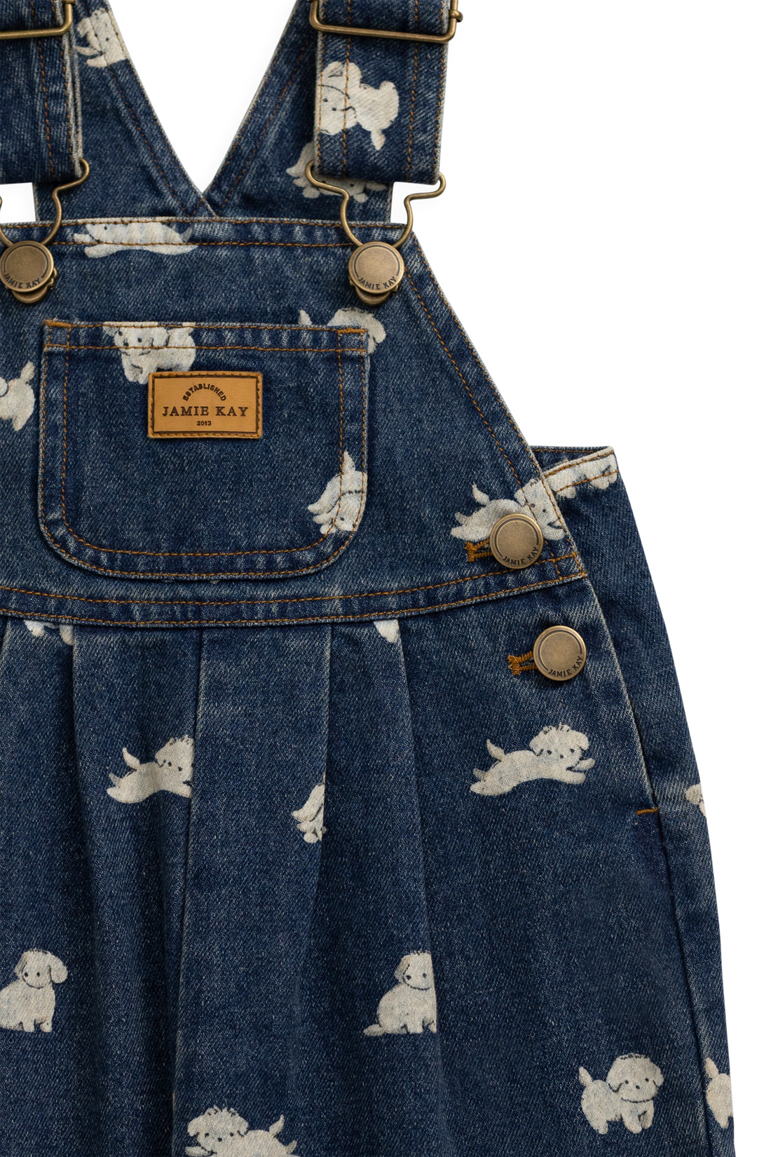 Close-up of a blue denim overall dress with a white puppy print, featuring brass-tone hardware and a tan Jamie Ray brand patch on the chest pocket.