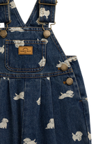 Close-up of a blue denim overall dress with a white puppy print, featuring brass-tone hardware and a tan Jamie Ray brand patch on the chest pocket.