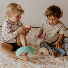 Two toddlers sitting on a patterned blanket playing with pastel crescent moon toys in a cozy indoor setting.