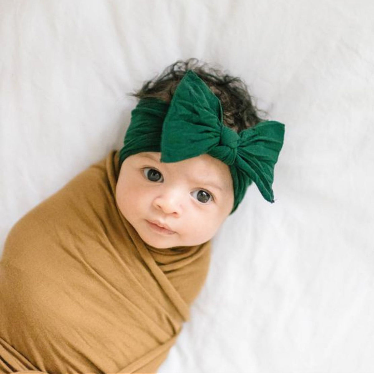 Baby wrapped in a mustard yellow blanket with a green headband on a white background