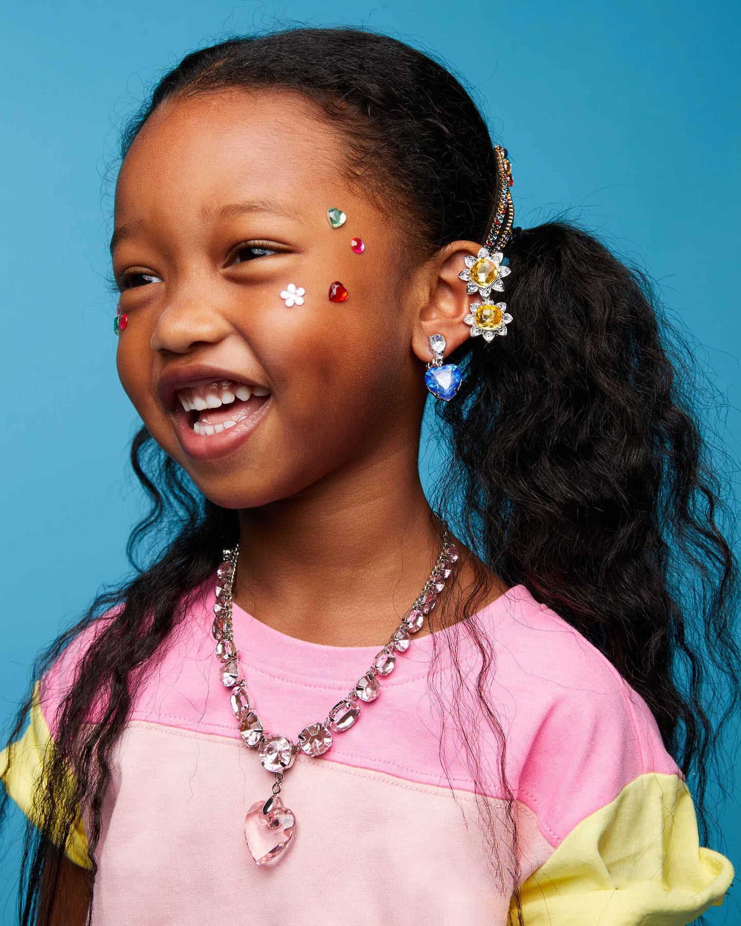 A young girl with dark curly hair in a ponytail smiles while wearing a gold daisy hair clip, a pink and yellow color-blocked shirt, a pink rhinestone necklace, and a blue rhinestone earring against a solid blue background.