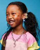 A young girl with dark curly hair in a ponytail smiles while wearing a gold daisy hair clip, a pink and yellow color-blocked shirt, a pink rhinestone necklace, and a blue rhinestone earring against a solid blue background.