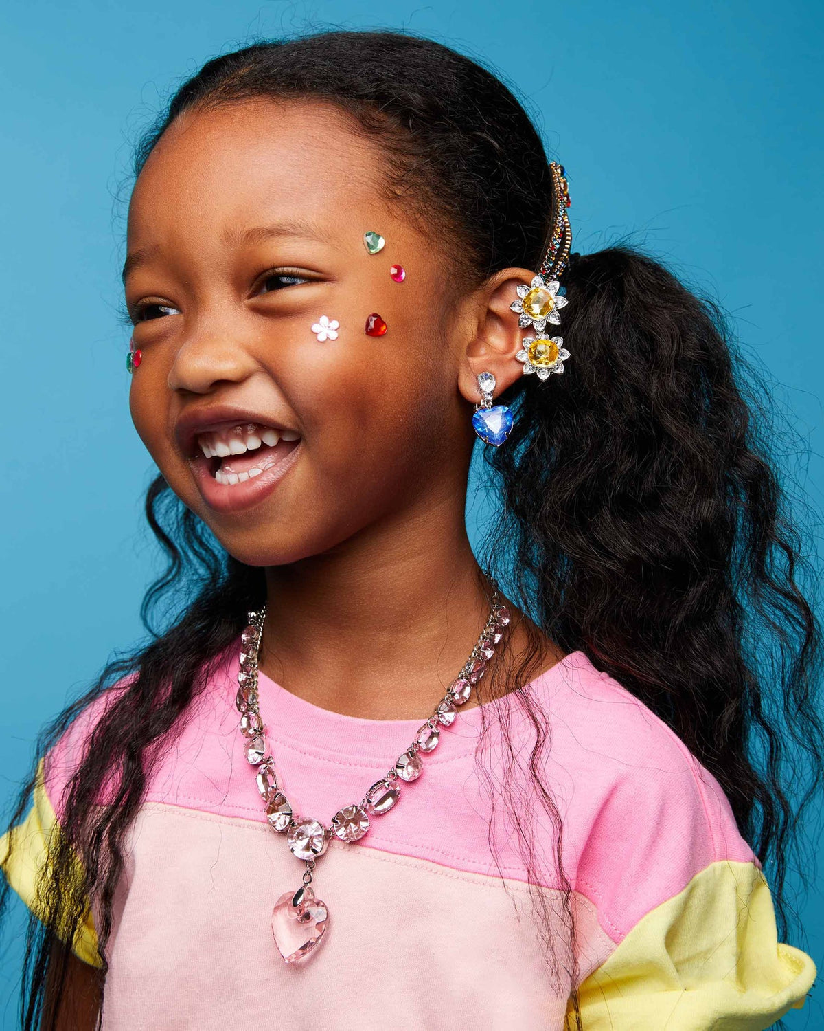 A young girl with dark curly hair in a ponytail smiles while wearing a gold daisy hair clip, a pink and yellow color-blocked shirt, a pink rhinestone necklace, and a blue rhinestone earring against a solid blue background.