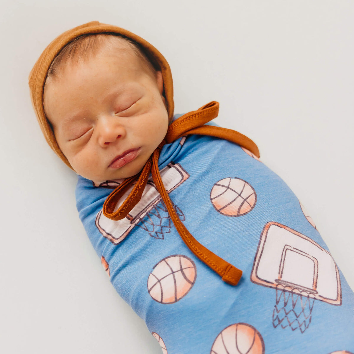 Newborn baby swaddled in a blue blanket with basketball patterns, wearing a brown bonnet.