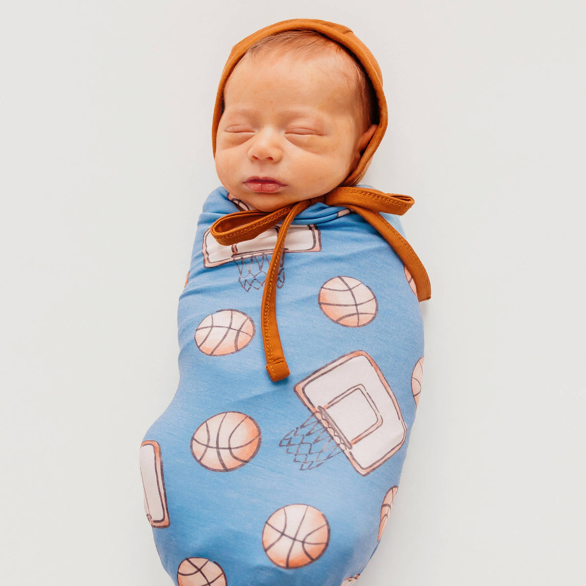 Newborn baby swaddled in a blue blanket with basketball patterns, wearing an orange bonnet.