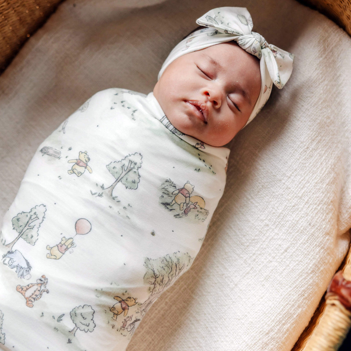 Newborn baby swaddled in a Winnie the Pooh patterned blanket with a headband on a soft surface.