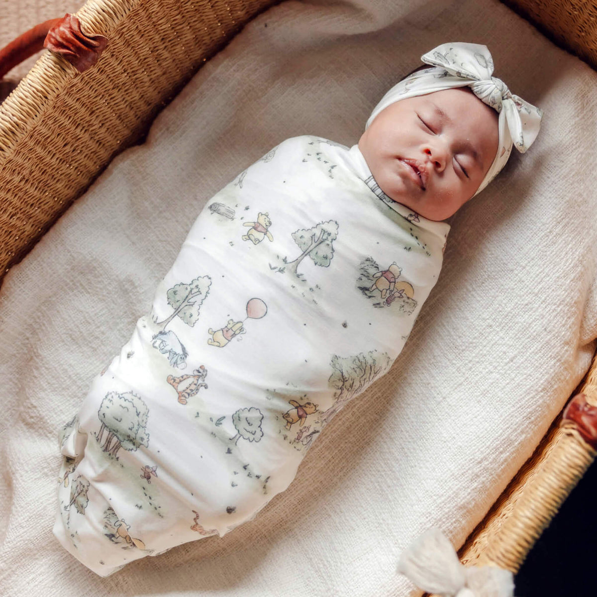 Newborn baby swaddled in a patterned blanket with a headband, lying in a woven basket.