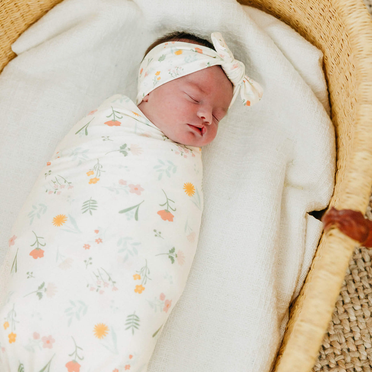 Newborn baby swaddled in a floral blanket with a headband, lying in a woven basket.