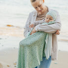 Woman holding a baby wrapped in a green patterned wrap on a beach.