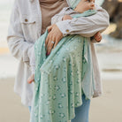 Person holding a baby wrapped in a green patterned wrap on a beach.