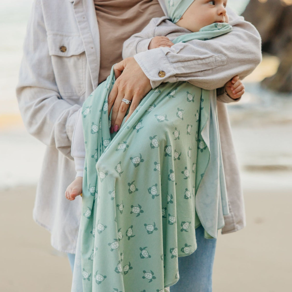 Person holding a baby wrapped in a green patterned wrap on a beach.