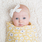 Baby wrapped in a yellow floral blanket with a white bow headband on a soft white background
