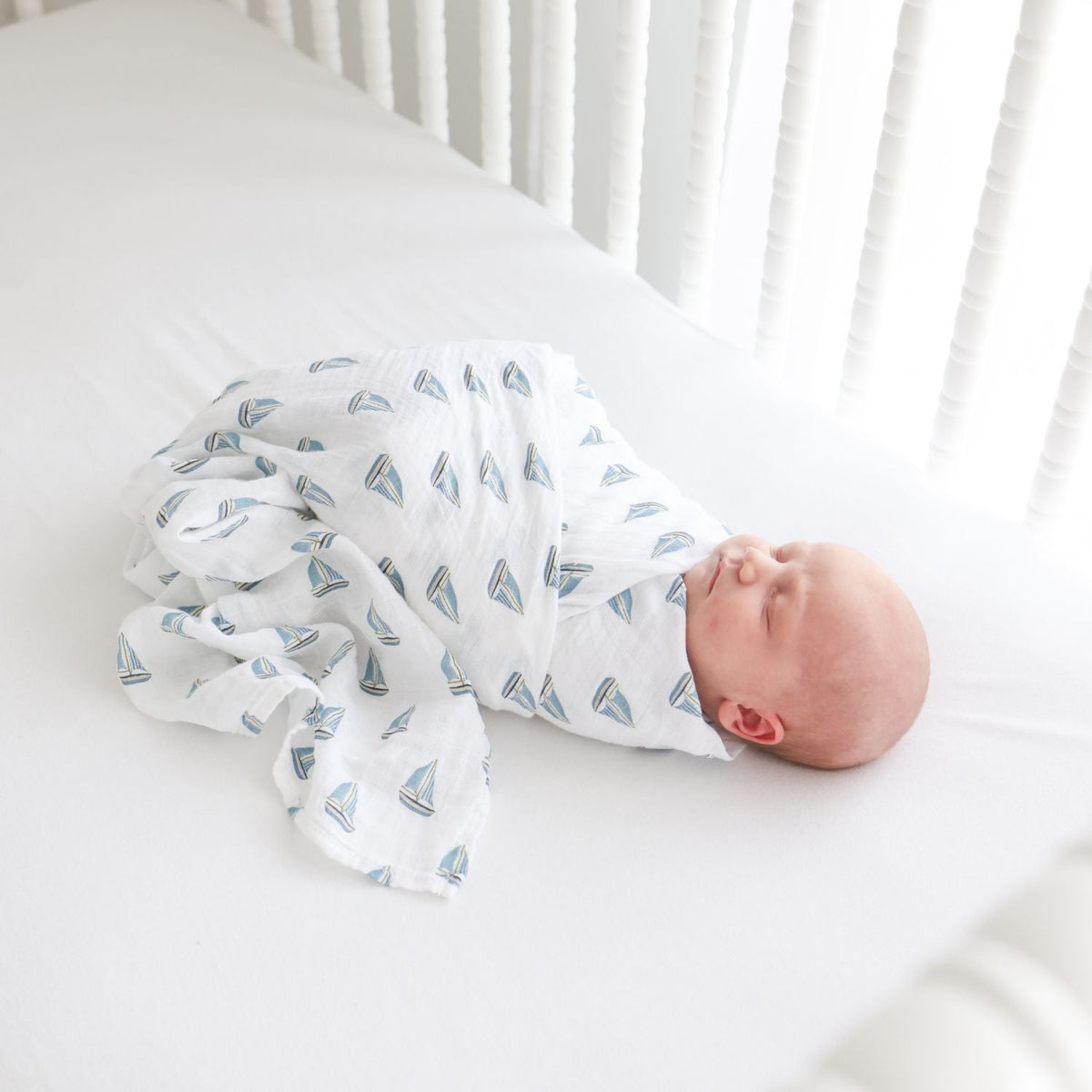 Newborn baby swaddled in a white blanket with blue sailboat pattern in a crib.