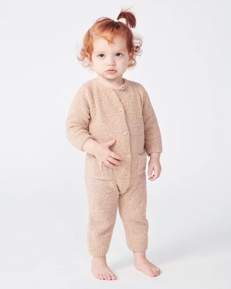 A toddler with reddish-brown hair stands facing forward, wearing a long-sleeve fuzzy romper in a warm pecan brown color by 7AM Enfant, against a plain white background.