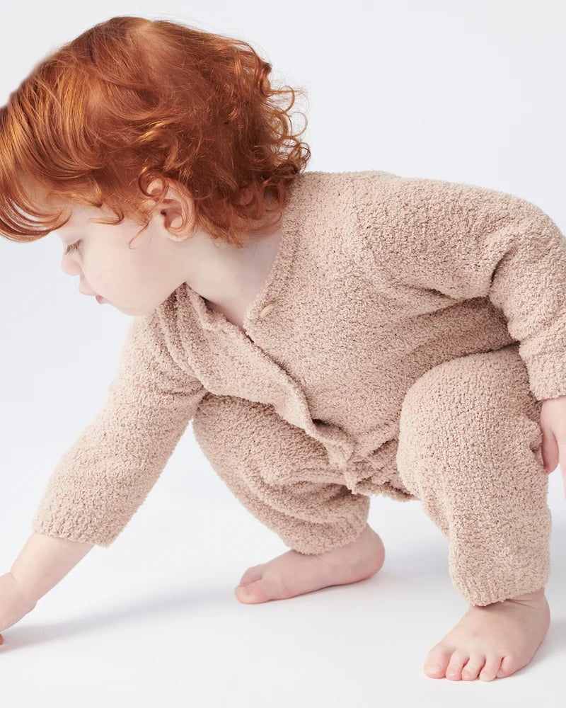 A toddler with reddish-brown hair is crawling on a white surface, wearing a long-sleeve fuzzy romper in a warm pecan brown color by 7AM Enfant.