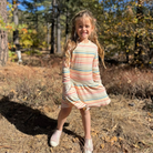 A young girl with long blonde hair stands outdoors in a wooded area wearing a long-sleeve striped dress with white sneakers.