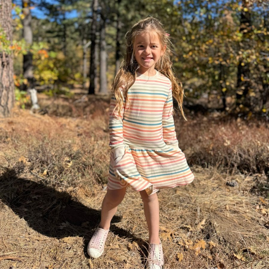 A young girl with long blonde hair stands outdoors in a wooded area wearing a long-sleeve striped dress with white sneakers.