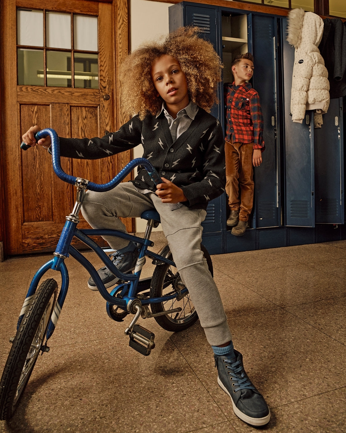 Child on a blue bicycle in a school hallway with lockers and another child in the background.